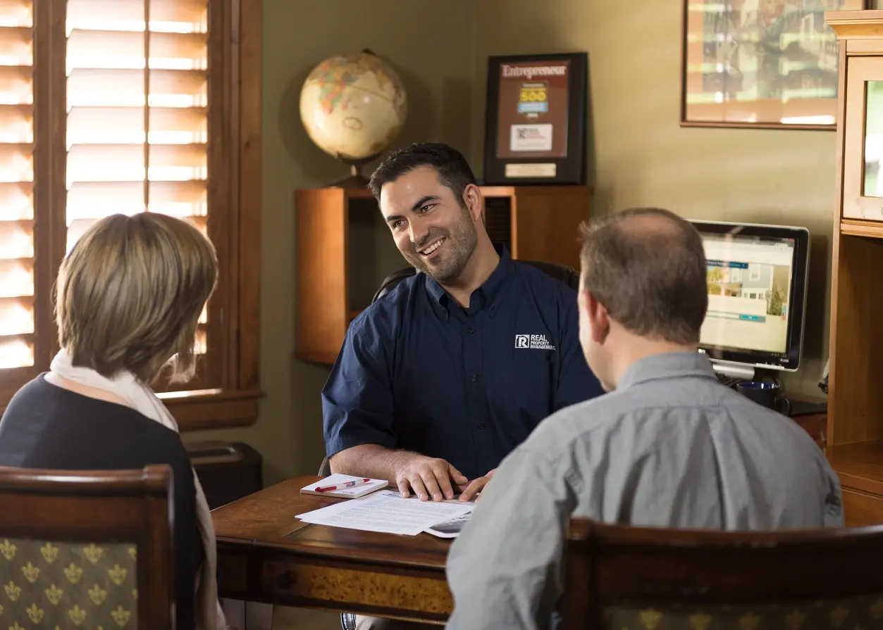 Real Property Management associate discussing lease with pair of customers seated at desk.